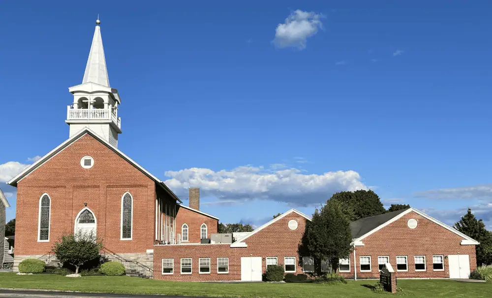 A picture of a red brick church set against a bright blue sky. The church has a white belfry and steeple and tall stained glass windows framing the white front door on the left side of the picture. To its right is a long one-story section with two peaked roofs. There are a few trees in the picture behind the church with green leaves in full canopies.