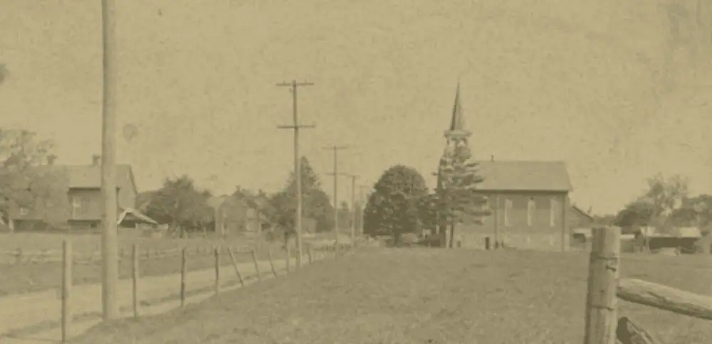 An undated sepia-toned photo showing the brick 1889 Schoeneck Moravian Church with no additions. Behind the church is what looks like the end of a barn and possibly a stables. In the foreground is a grassy field. There is a fence by the dirt road with telephone poles, indicating electricity is available. Across the road is a farmhouse and a large barn.