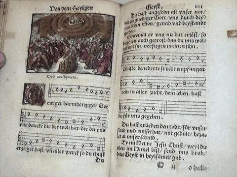 A close-up photo of an open edition of the 1544 German Hymnal. There is a large illustration of people in worship at the top of the left-hand page. Underneath is a hymn with three tune stanzas with the words underneath each stanza. At the top of the right-hand page are two brief paragraphs. In the middle are three tune stanzas with the words underneath each stanza. At the bottom are another two small paragraphs. All of the text is in German.