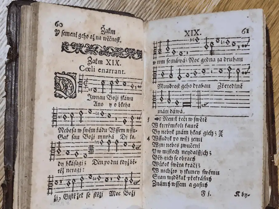 A close-up photo of a small black hymnal, open to pages 60 and 61 featuring Psalm 19. On the left-hand page is the beginning of a hymn with four tune stanzas, each with two verses written below. The hymn continues on the right-hand page with three more tune stanzas, each with just one verse underneath, possibly indicating a refrain. Underneath that hymn are words to possibly another verse. All of the text is in Czech.