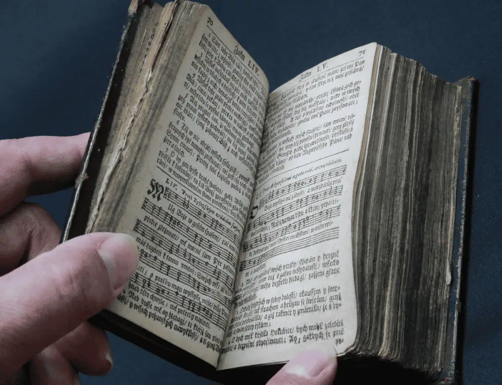 A close-up photo of a hand holding a very small hymnal, opened to show Psalm 54 on the left-hand page and Psalm 55 on the right-hand page. The left page has six small paragraphs followed by five tune stanzas with the words underneath each stanza. On the right page is three paragraphs at the top, four tune stanzas with the words underneath in the middle, and three paragraphs at the bottom. The text is written in Czech.