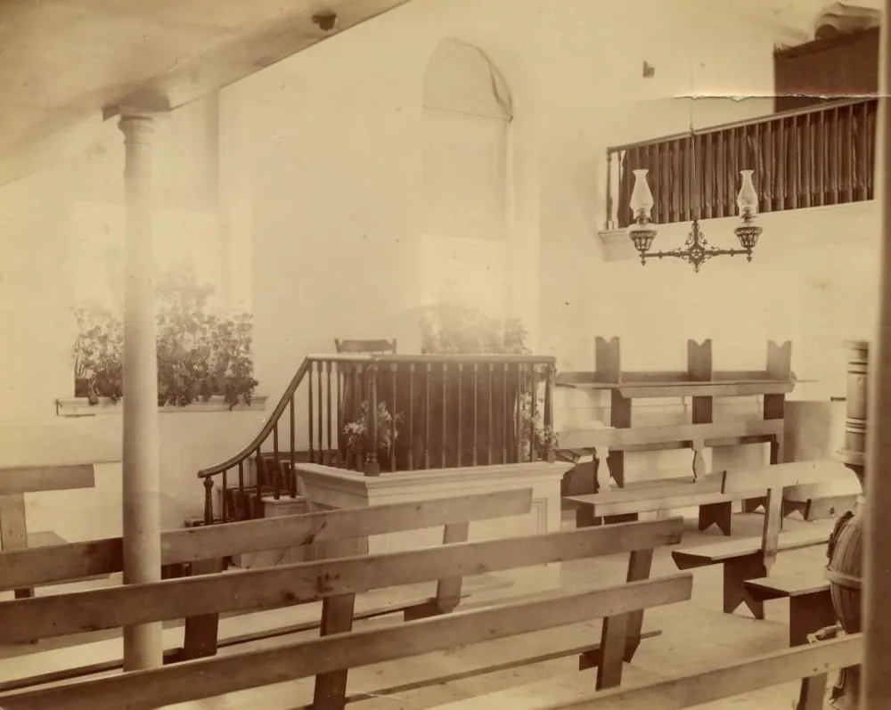 A sepia-toned photo of the interior of the 1792 church, circa 1887. An elevated railed-in communion table is in the center of the picture. There is a balcony visible on the left side of the picture, and supporting columns for a new balcony on the right.