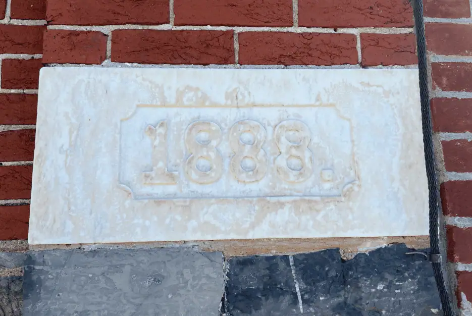 A color photo of the 1888 cornerstone of the building. The cornerstone is white with grey and black stones of the foundation beneath it. It is surrounded by red bricks.