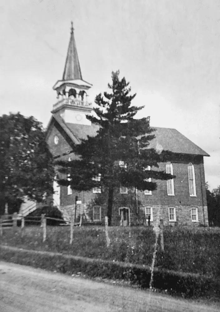 A black and white photo of the current red brick church building, as originally built in 1888-1889. You can see the church steeple, but the front of the church is obscured by two trees. Along the side of the building are five tall windows for the sanctuary with five smaller windows underneath for the classroom level.
