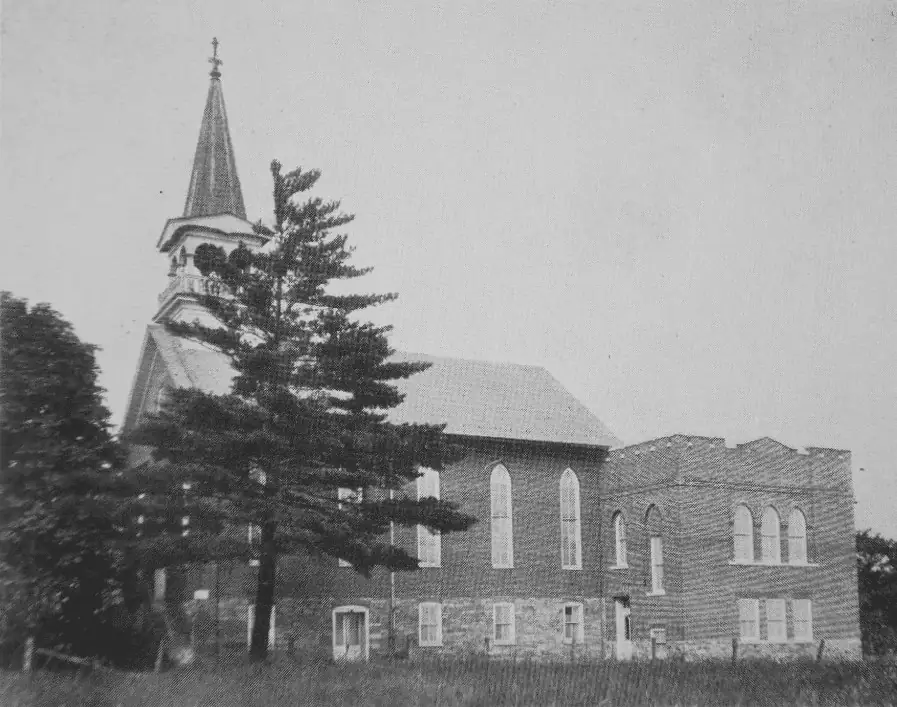 A black and white photo showing the 1889 brick church and the Sunday School addition. The addition is square and and there is a door in front. The windows on the first floor are rectangular and the windows on the second floor are taller and rounded at the top. They match the size and shape of the windows on the church. The addition has decorative castle-like details along the top of the building with a flat roof.