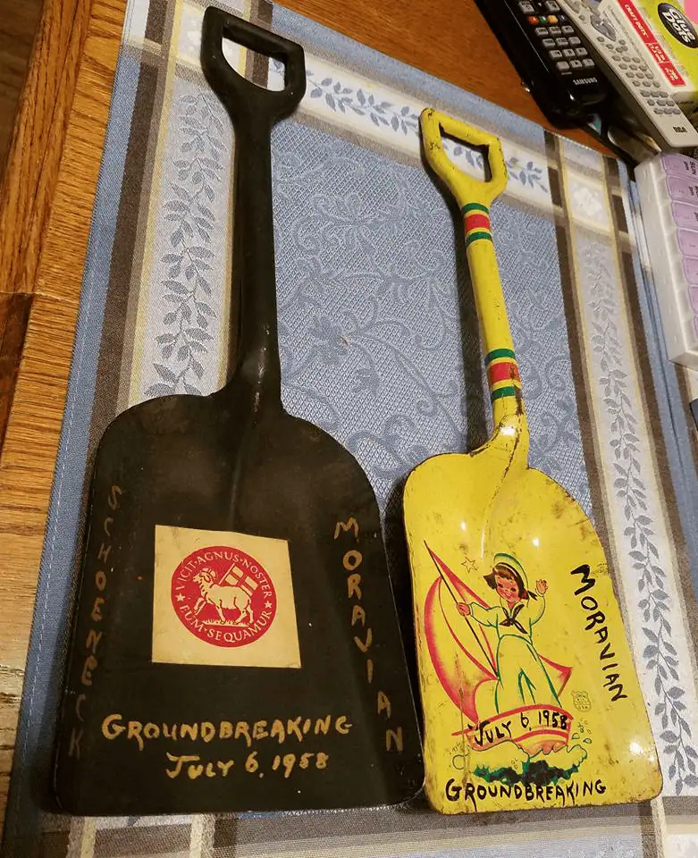 Two ceremonial shovels used in the groundbreaking ceremony for the new Christian Education wing on July 6, 1958. The shovel on the left is black. On the blade is a red Moravian Seal in the middle with Schoeneck written on the left side and Moravian written in gold paint. The shovel on the right is yellow and features a young girl in a sailor suit standing on a sailboat on the blade. The words "July 6, 1958 Groundbreaking" are written on the bottom edge of the blade, with Schoeneck and Moravian on the sides.
