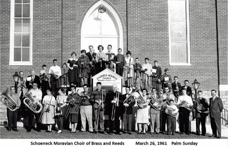 A black and white photo labeled "Schoeneck Moravian Choir of Brass and Reeds, March 26, 1961, Palm Sunday" Men and women are gathered in front of the church, standing on the steps and in front, surrounding the church sign. Some people are holding various brass instrument like tubas, trumpets, French horns, euphoniums, and trombones. Others are holding clarinets and oboes.