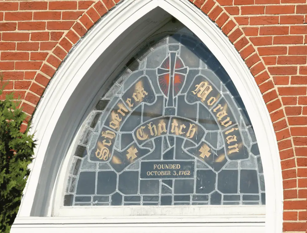 A color photo of the stained glass window above the front doors. The words Schoeneck Moravian Church are written in gold script with Founded October 3, 1762 in smaller gold letters.