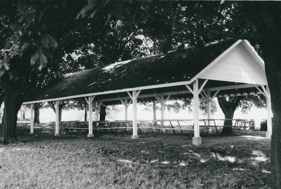 A black and white picture of the church pavilion from June 1978. The pavilion is white with a dark shingle roof with a number of picnic tables inside. The pavilion is surrounded by grass and trees.