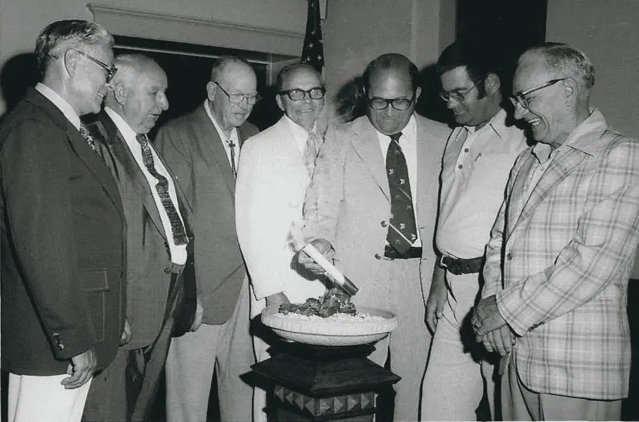 In this black and white photo, seven smiling men in suits stand around a white bowl filled with sand atop a brown church stand. One of the men holds a rolled paper, which is burning from the bottom end, over the bowl.