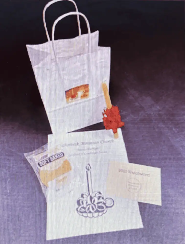 A color photo of a 2020 Christmas Eve To-Go bag and its contents. There is a small white bag with white handles, with a red-frill wrapped beeswax candle, a packaged cookie which says "soft baked", a small card which reads "2021 Watchword", and an ode for Schoeneck's Christmas Eve Lovefeast and Candlelight Service which has a clip-art image of a lit, frilled candle in a holder on it.