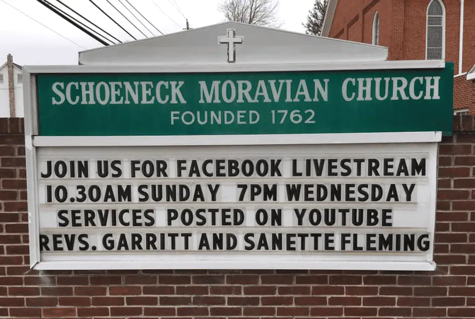 A close-up picture of the church sign. The sign enclosure is made of red bricks. at the top is an acrylic green sign which reads "Schoeneck Moravian Church, Founded 1762". Beneath the green sign is a place for individual letters to be placed in four white rows. The letters are black and the message reads "Join us for Facebook Livestream 10:30 am Sunday, 7pm Wednesday. Services posted on YouTube. Revs. Garritt and Sanette Fleming."