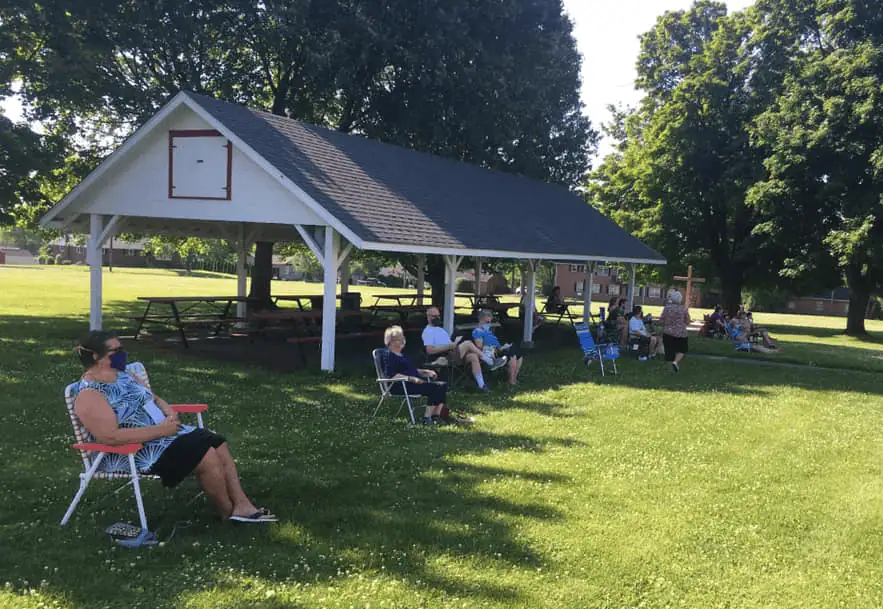A color photo of the grove and pavilion on a sunny day. There are a number of people sitting in lawn chairs with several feet in between chair. Each person is wearing a protective mask covering their mouth and nose.