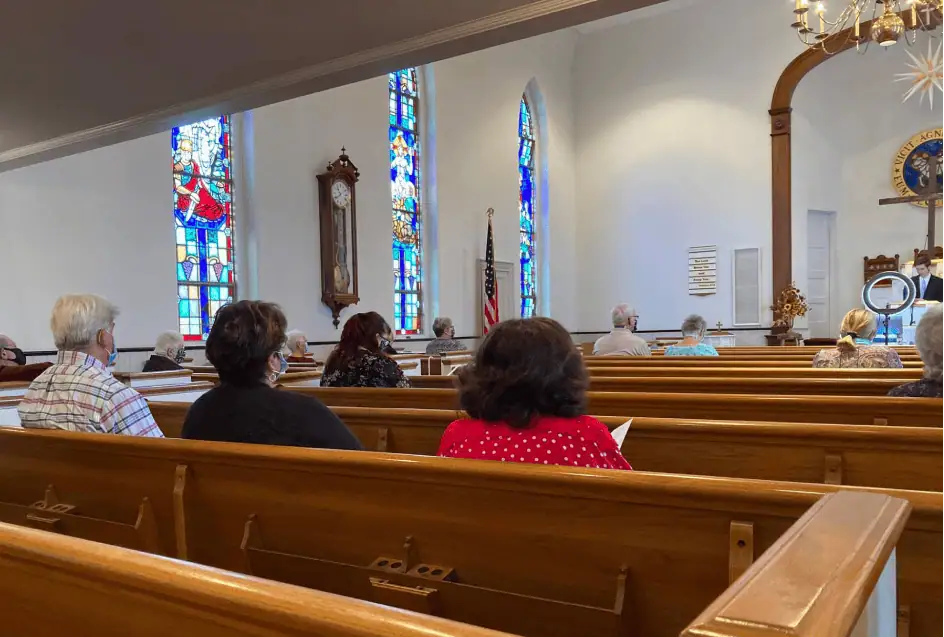 A color photo of the inside of the church sanctuary. There are a number of worshippers sitting in the pews. They are spread apart by an empty pew between congregants. They are all wearing protective masks covering their mouth and nose.