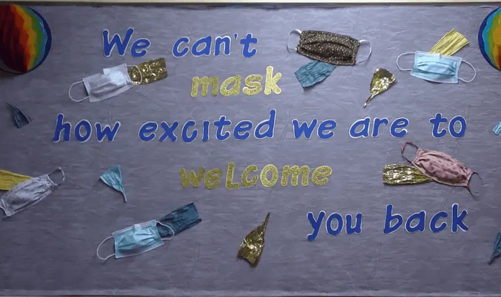 A color photo of a bulletin board with several different styles of protective face masks attached to it. The words read: We can't mask how excited we are to welcome you back."