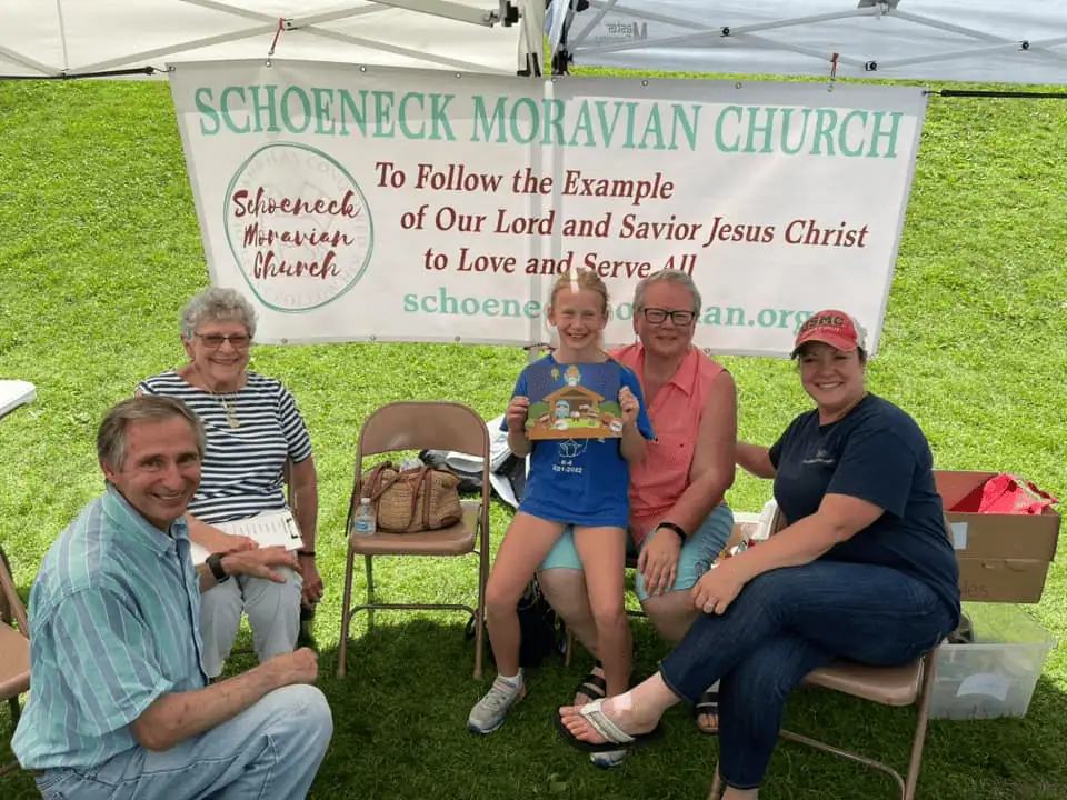 An intergenerational group of 5 Schoeneckians sitting under the Schoeneck Moravian Church tent at a Nazareth Day celebration in Nazareth Borough Park. There is a banner behind them with our Mission Statement: To follow the example of our Lord and Savior Jesus Christ to Love and Serve All.