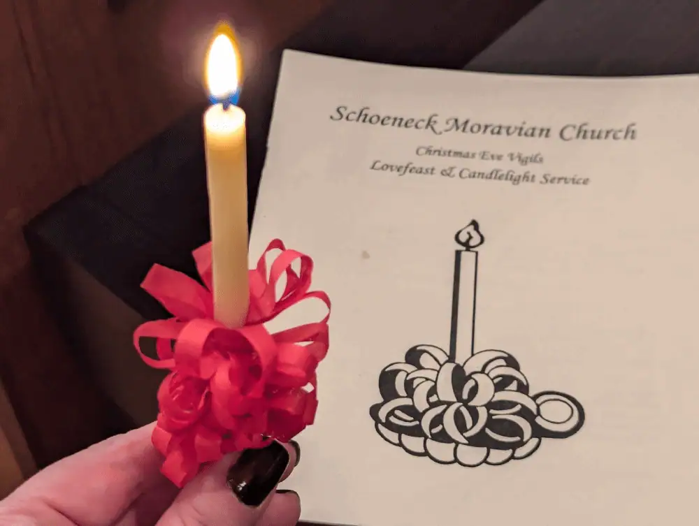 A woman's hand holds a lit red-wrapped beeswax candle in front of one of Schoeneck's Christmas Eve Lovefeast odes.