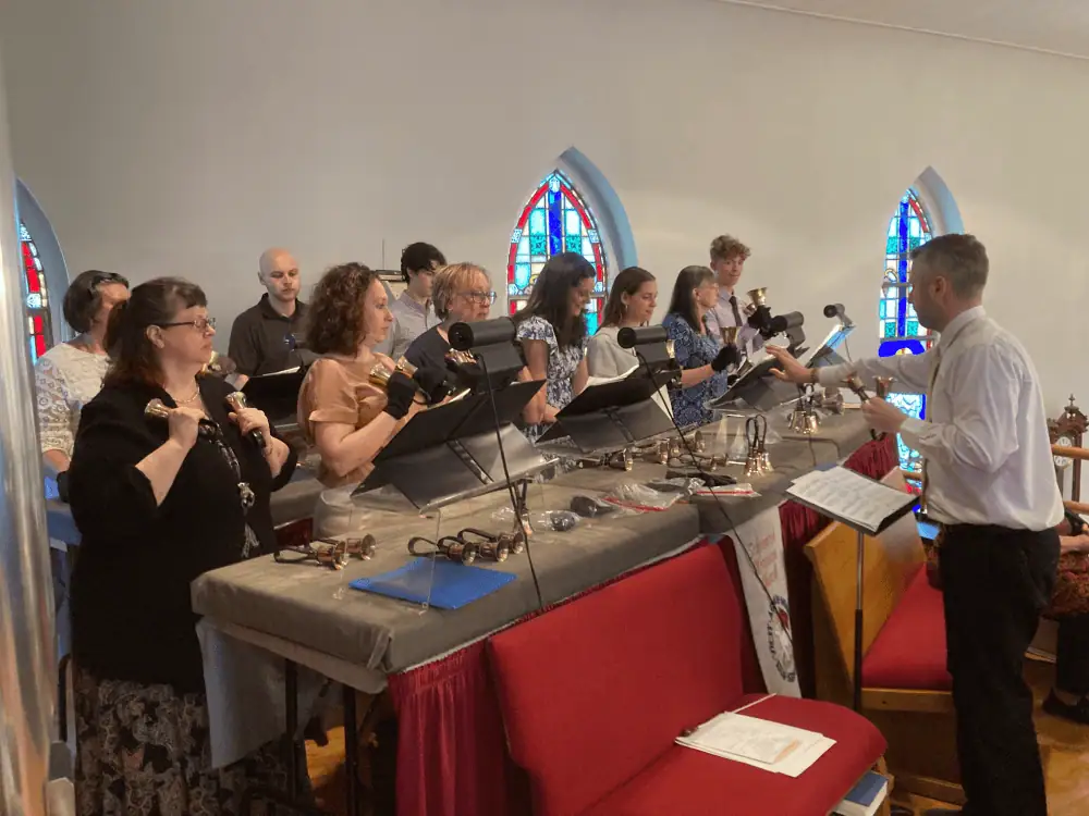 A photo of the Senior Bell Choir playing at the bell tables in the balcony during a worship service with our Director of Music Ministries directing. The choir is made up of men and women of various ages. The bell tables have a grey cushion with burgundy table skirts. The bells are golden with black handles.