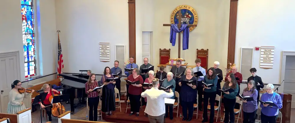 A photo of the Senior Choir's 2025 Lenten Cantata. The choir is assembled on the raised pulpit area, men in the back and women in the front. In front of the choir is our Director of Music Ministries, Ryan Morrow, directing. On the left side of the picture is a young women, standing as she plays a violin. To her left is an older woman, sitting as she plays the viola. The black grand piano is in the corner with a woman playing it. In the pulpit is a cross draped with purple-blue fabric.