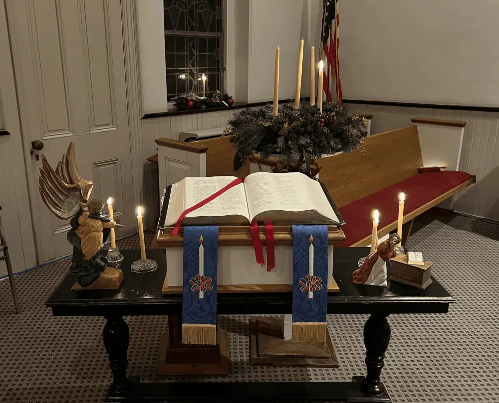 The photo is of part of the sanctuary decorated for the Advent Candlelight Holy Communion Service. There is a dark brown table which has the small white pulpit lectern on it. The lectern holds the Holy Bible, with blue Advent paraments. The paraments have a lit candle wrapped with red and green frills on them. On the left side of the Bible is the Angel Gabriel from Schoeneck's putz with two lit candles behind it. On the right of the Bible is the figure of Mary from the Annunciation, also from our putz, with two lit candles behind her. Behind the table is Schoeneck's Advent wreath, with one lit beeswax candle.