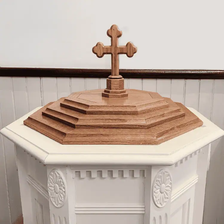 A photo of Schoeneck's baptismal font. It is hexagonal in shape, with a white base. The top is a medium warm wood with a cross on top.
