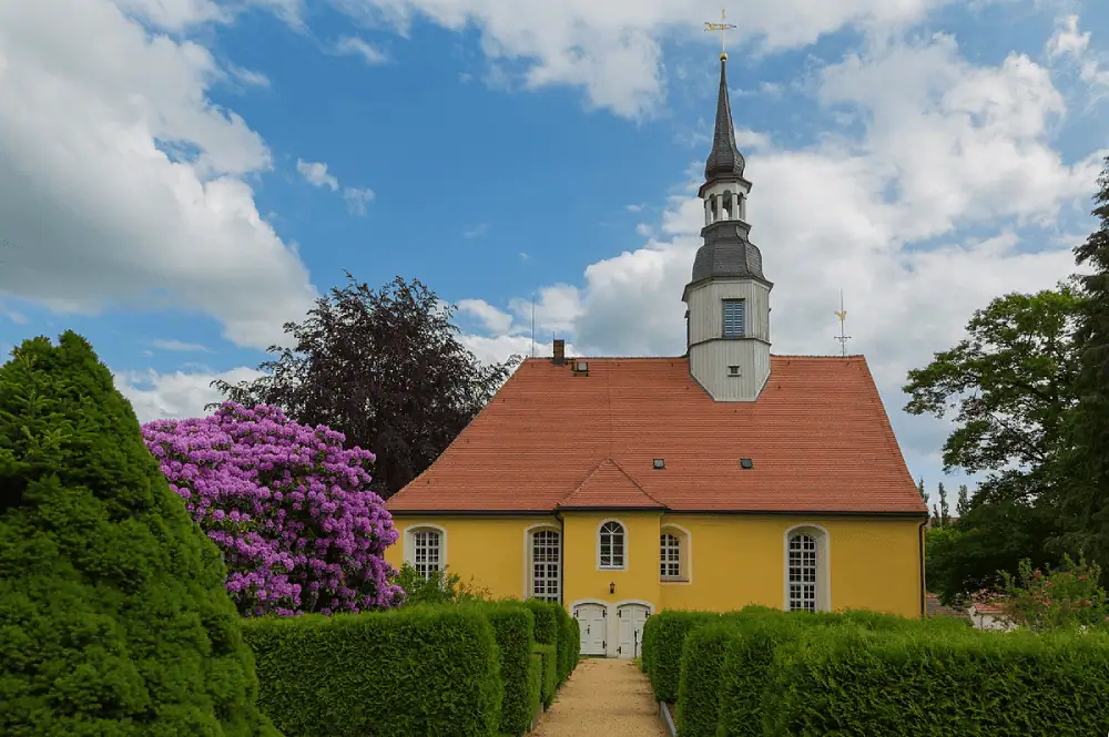 Under a bright blue sky with puffy white clouds stands a small church. There is a white steeple bell tower on the right side of the roof. The roof itself is a burnt orange color, and the walls are a bright yellow. There are large leafy trees behind the church. In front of the church to the left is a large bush with purple flowers. There are hedges on both sides of a path leading to the church doors.
