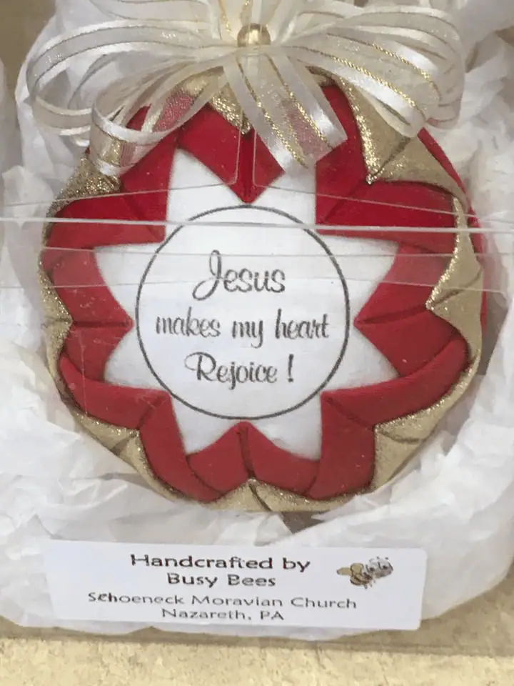 A round red, white, and gold fabric ornament with "Jesus makes my heart Rejoice!" in a thin black circle on the front. The ornament is in a clear, plastic square box with white tissue paper on the bottom. A sticker on the box reads "Handcrafted by Busy Bees. Schoeneck Moravian Church, Nazareth, PA."