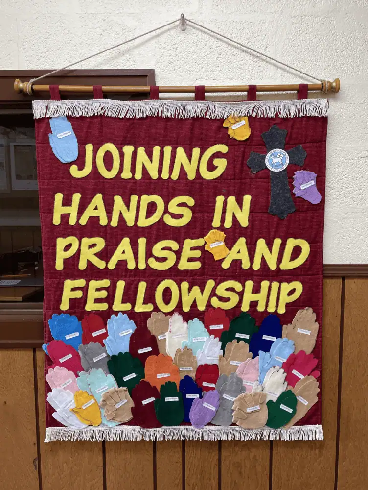 A banner made by the Christian Education Ministry that reads "Joining Hands In Praise and Fellowship". There are felt cutouts of hands of different sizes and colors filling the banner underneath the words.
