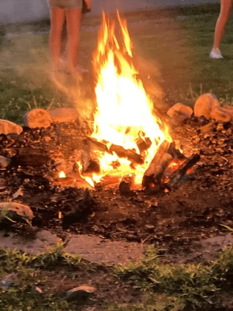 A close-up photo of a campfire at Camp Hope features the traditional circle made of rocks around a roaring fire reaching to the night sky with stacked wood visible in the middle of it. The fire circle is surrounded by dirt and grass.