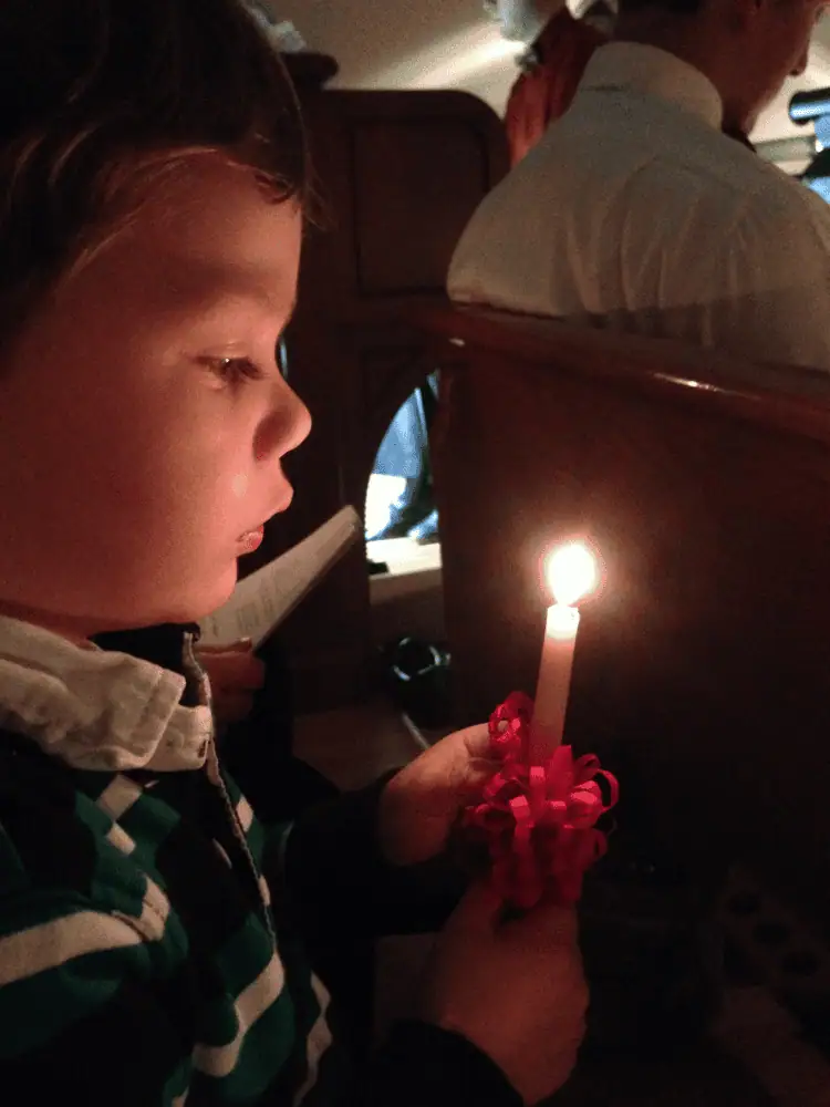 A young boy holds a lit red-wrapped beeswax candle on Christmas Eve. He is gazing at the flame, and his face is lit by candlelight.