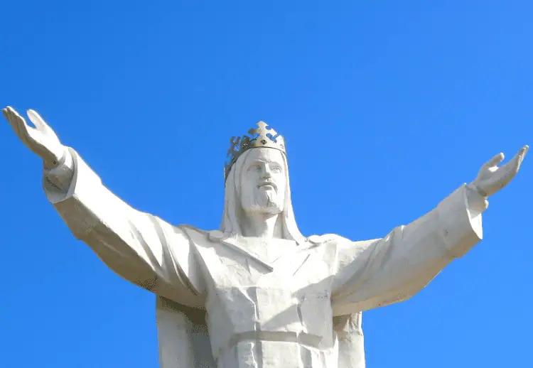 Against a bright blue sky, the top half of the Christ the King monument reaches towards your with outspread hands. The statue is white and Christ wears a golden crown.