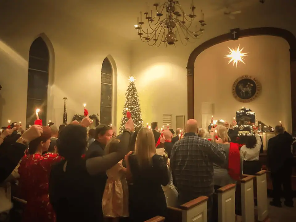 A picture of the Schoeneck congregation lifting their lit red-wrapped candles in the air. The glowing Christmas tree is in the just left of center of the top of the photo, and the shining Moravian Star above our Putz is just right of center.