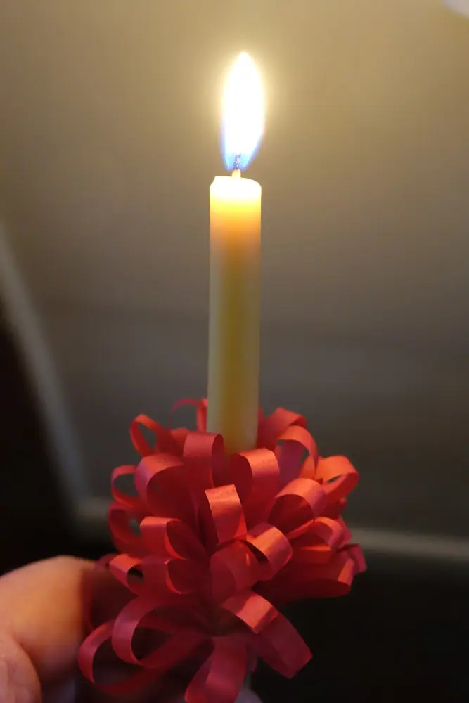 A close up photo of someone holding a red-wrapped beeswax candle. The paper frill is full of curls, the candle is a light-golden yellow, and the flame shines brightly.