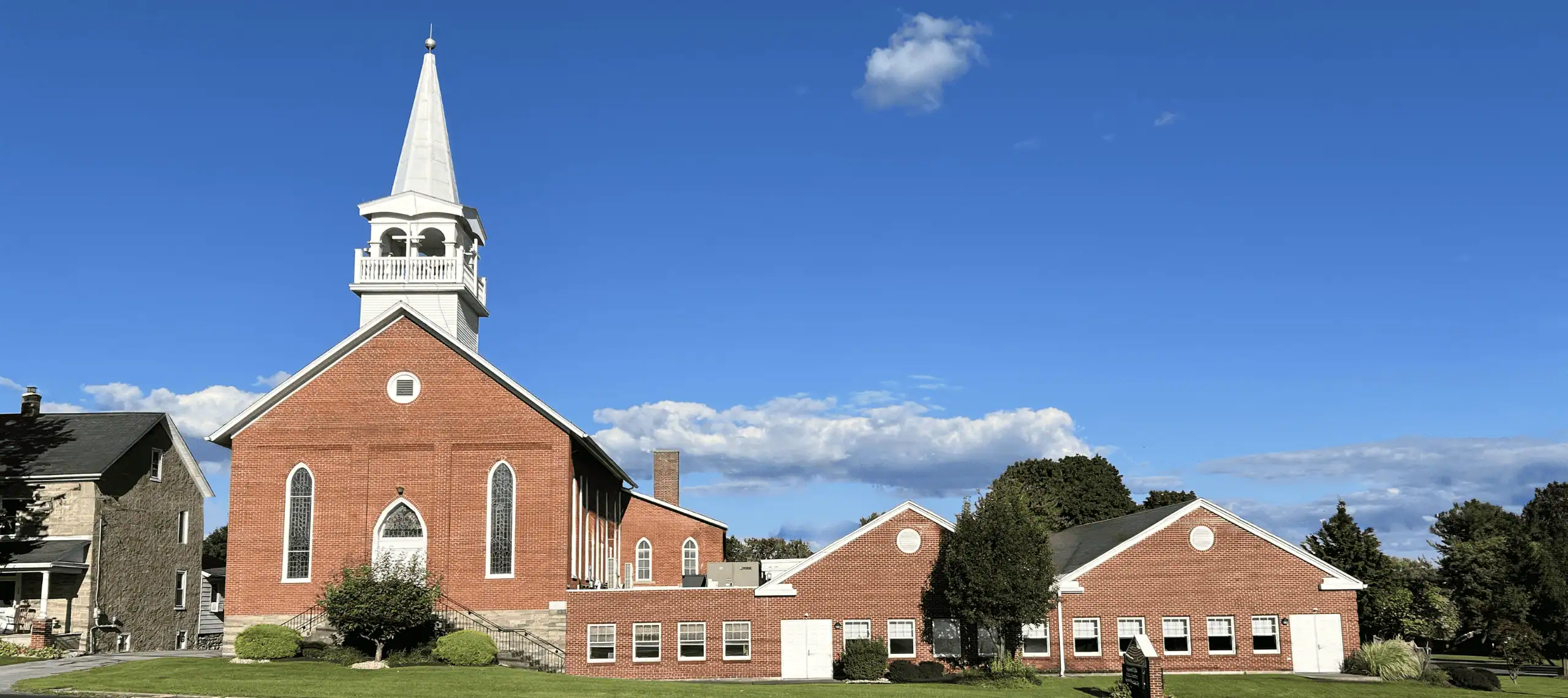 A picture of a red brick church set against a bright blue sky. The church has a white belfry and steeple and tall stained glass windows framing the white front door on the left side of the picture. To its right is a long one-story section with two peaked roofs. There are a few trees in the picture behind the church with green leaves in full canopies.