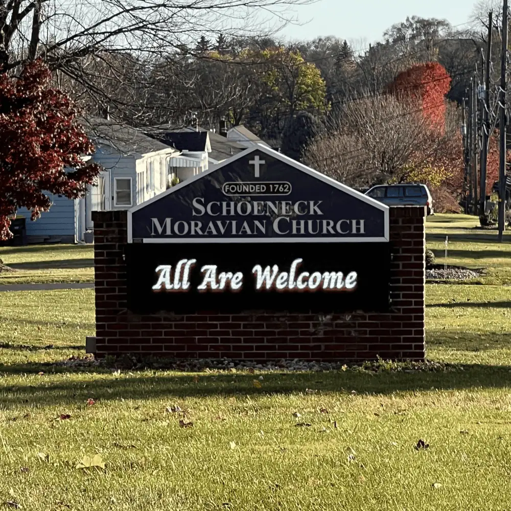 A picture of the church sign on the front lawn. It has a brick surround with a LED sign. On top the sign says Schoneck Moravian Church - Founded 1762. On the LED sign is the message "All Are Welcome!"