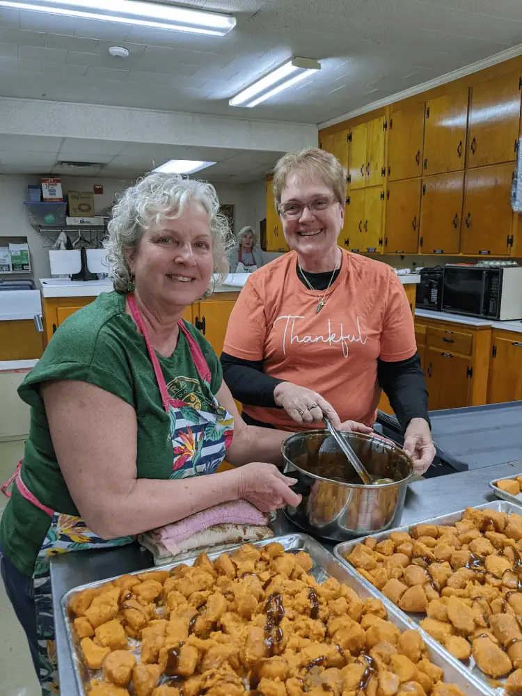 Two women stand in front of two large stainless steel trays filled with sweet potatoes, preparing to drizzle a glaze over them before cooking.