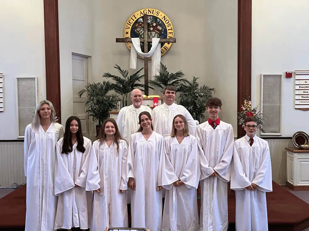 Seven youth wearing white robes stand in front of Rev. Otto Dreydoppel, Jr. and Rev. Ian Edwards, both wearing their surplices. The pulpit is decorated with palms plants, and a cross with a white cloth draped over it.