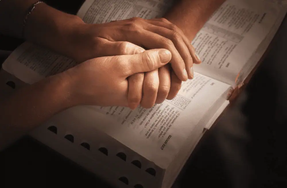 A close-up image of two people clasping hands over an open Bible.