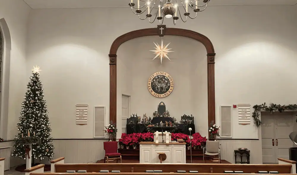 A picture of Schoeneck's sanctuary decorated for Christmas Eve services. The Christmas tree glows with white lights, with multicolored Moravian stars scattered among its branches, and a Moravian star on top. The Advent Wreath stands in front of the tree, toward the pews. At the top of the pulpit area is Schoeneck's Moravian Star, with white and yellow points. The pulpit itself features Schoeneck's putz of hand-carved wooden figures. Below the putz are a bank of poinsettias. Over the front sanctuary doors are a garland of winter greens with golden bows, pine cones, and red berries.