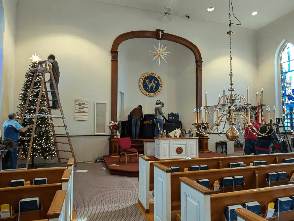 A view of the front of the sanctuary. On the left is an artificial Christmas tree. Next to it is a ladder. A man holds the ladder while a youth adjusts the branches at the top of the tree. In the middle, two people are arranging Schoeneck's putz in the pulpit. There is a chandelier hanging just above the pews, ready to be cleaned. On the right are two people with a ladder.
