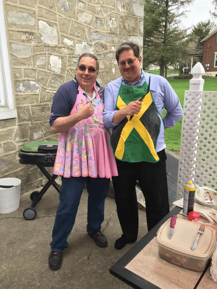 Two Schoeneckian men pose for a silly picture as they prepare food for a fellowship meal. They are wearing frilly aprons and posing with grilling instruments pointing at each other like swords.