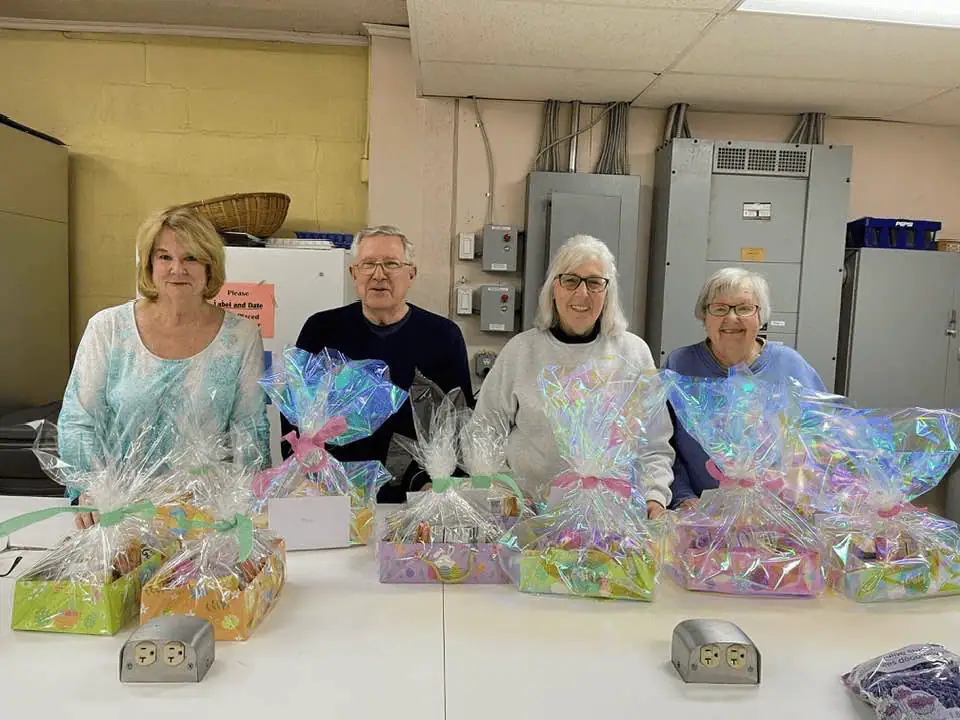 Three women and a man stand behind the white prep and serve island in Schoeneck's kitchen. In front of them are several easter packages wrapped in iridescent cellophane with colorful bows.