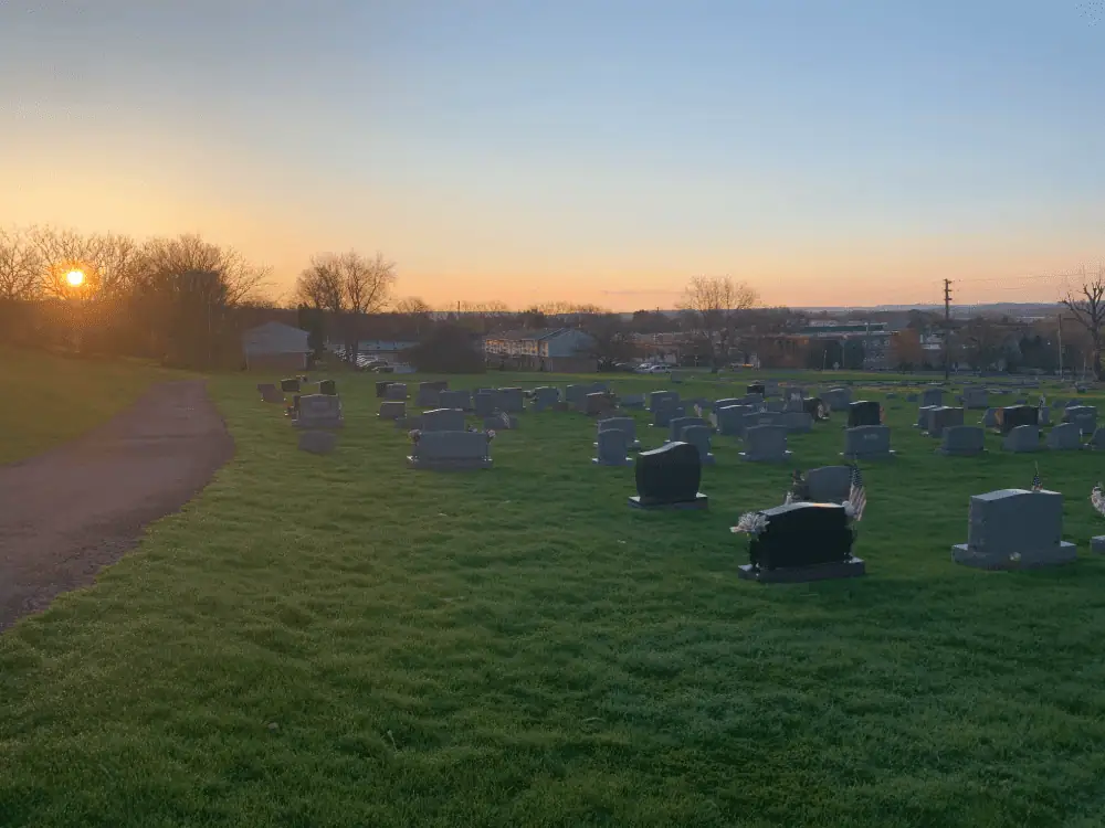 The sun is rising through the trees overlooking Schoeneck's cemetery, God's Acre. The sky is shades of orange and yellow as it fades upwards to blue. On the left is the macadam path, with numerous gravestones amid the green grass on the right of the image.