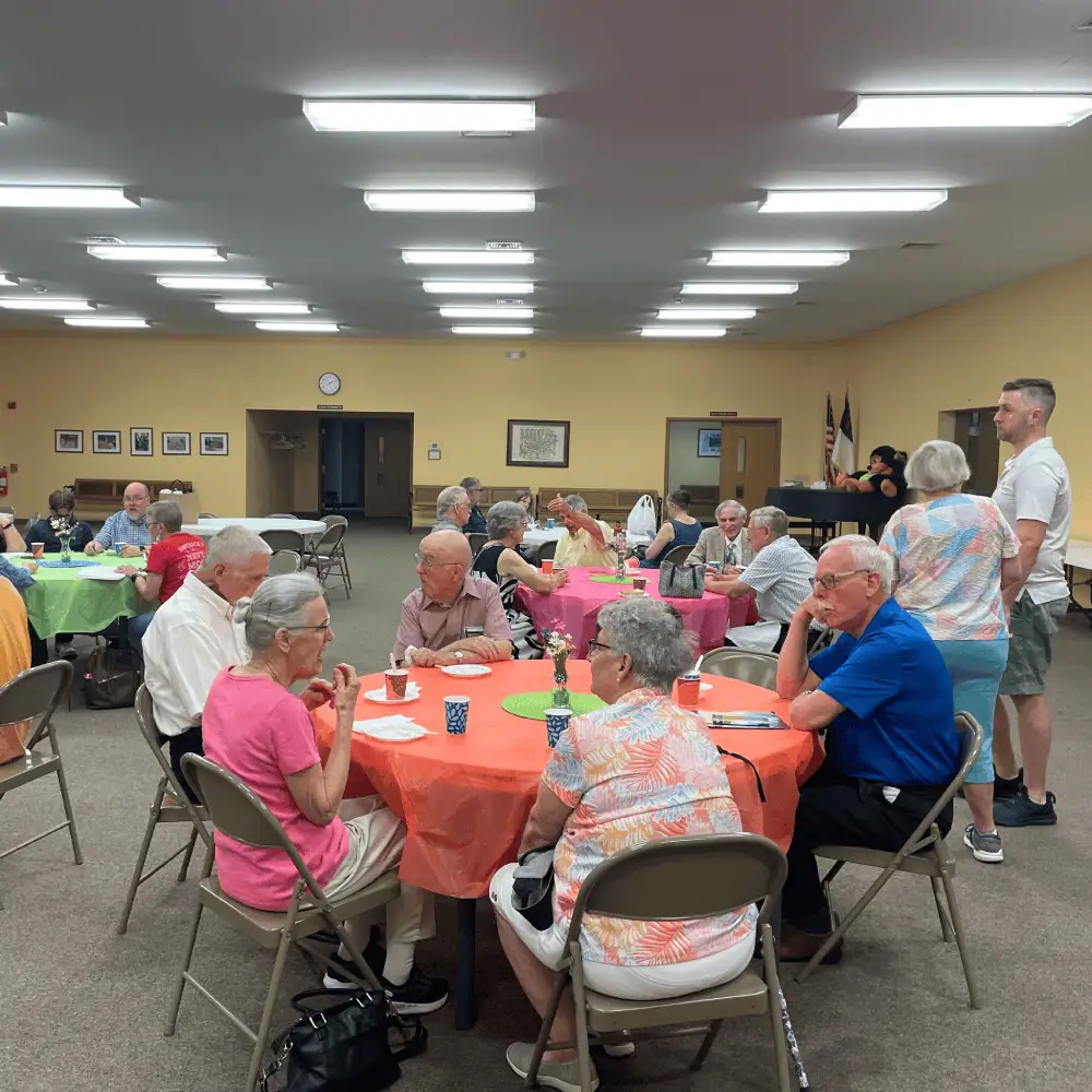 A photo of a Snack 'N Yak fellowship. There are several round tables in the Fellowship Hall decorated with brightly colored plastic tablecloths. Many Schoeneckians - young and old and in between - sit around the tables, enjoying snacks, beverages, and conversation.