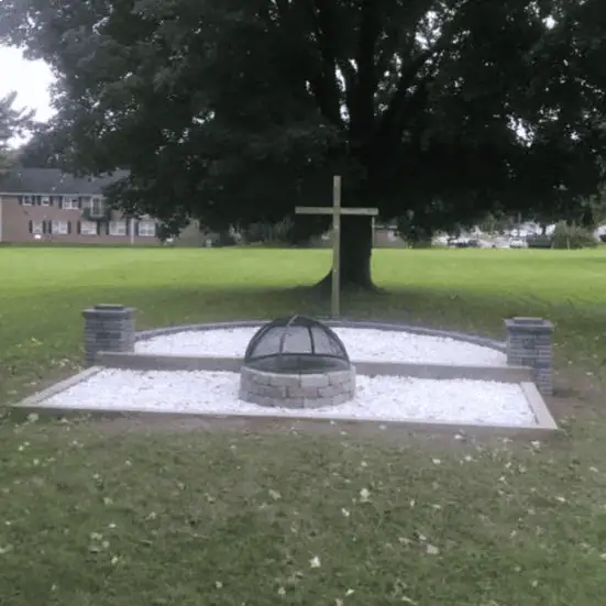 The fire pit at Schoeneck. There are two areas - a rectangular area with white stone which has a circular firepit made of grey bricks with a screen dome cover in the middle. The other area is slightly raised and curved, with white stone and a wooden cross standing at the end. Two grey stone pillars stand at the side edges at the intersection of the two areas.
