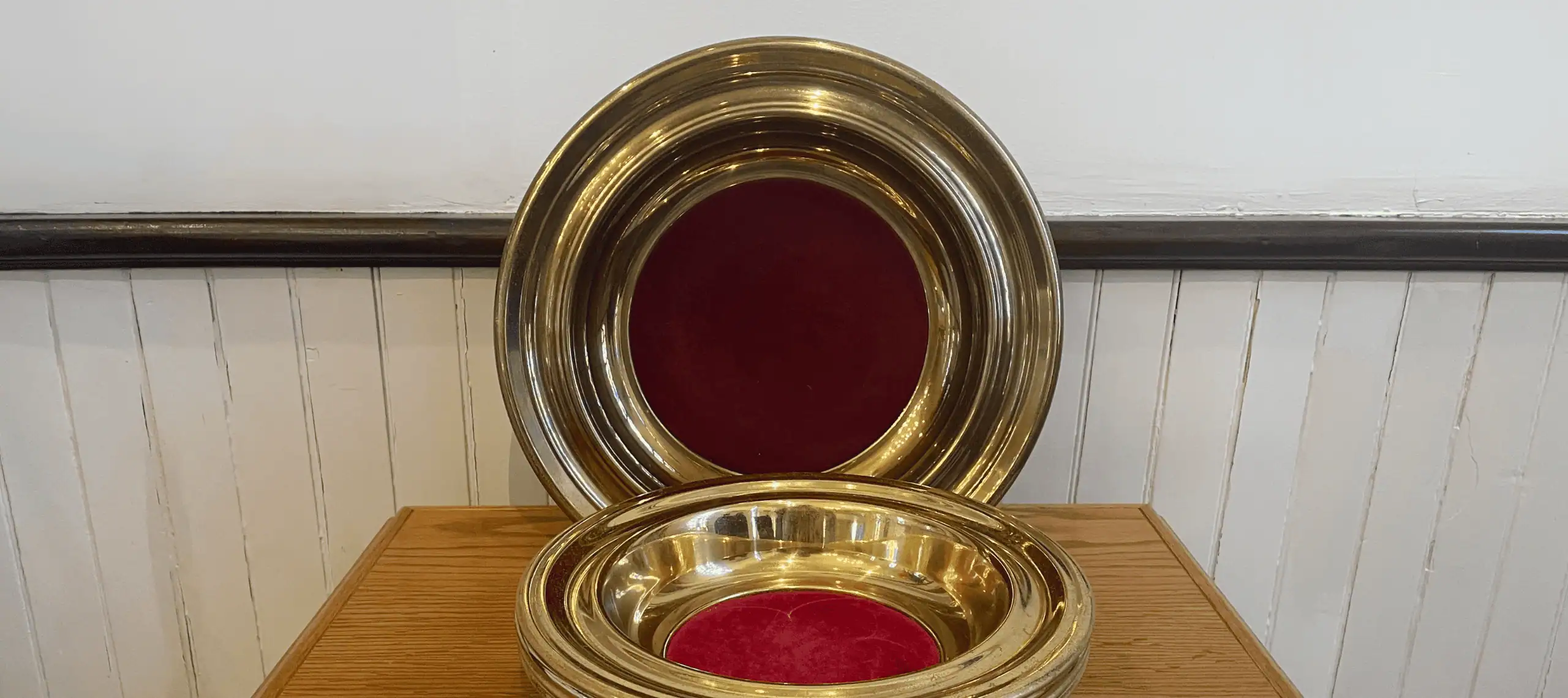 Schoeneck's gold offering plates are stacked together on a medium wooden tabletop in front of a larger offering plate which is standing against the white wainscoting of the wall.