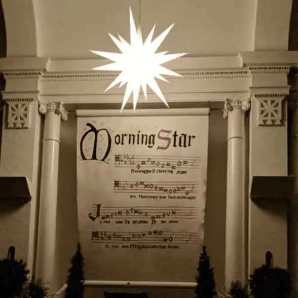 A photo of a brightly glowing Moravian star above a banner featuring the first verse of "Morning Star, O Cheering Sight" in the sanctuary of Gnaddenhutten Moravian Church in Gnaddenhutten, Ohio.