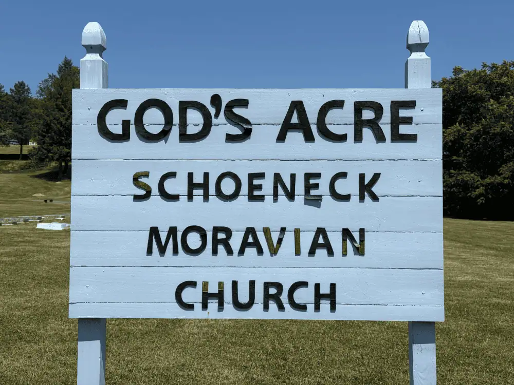 The sign for Schoeneck's cemetery on a sunny day. The sign is made of wood slats that have been painted grey. The words are wood as well and are black: God's Acre - Schoeneck Moravian Church.