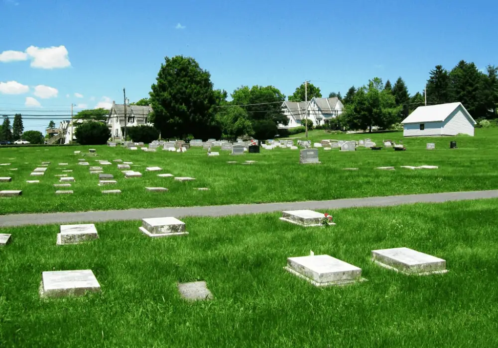 A color picture of the Schoeneck cemetery, known as God's Acre. The sky is a bright sunny blue and the grass is a verdant green. There are flat white markers at the bottom of the picture. Just above the markers is a gravel road. At the top of the image are more flat white markers.