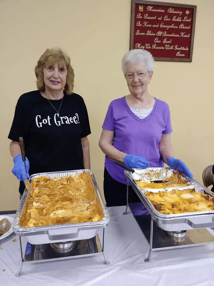 Two older Schoeneckian women stand behind a table with buffet servers ready to dish up their delicious contents. One of the women is wearing a black t-shirt with white lettering that reads "Got Grace?"